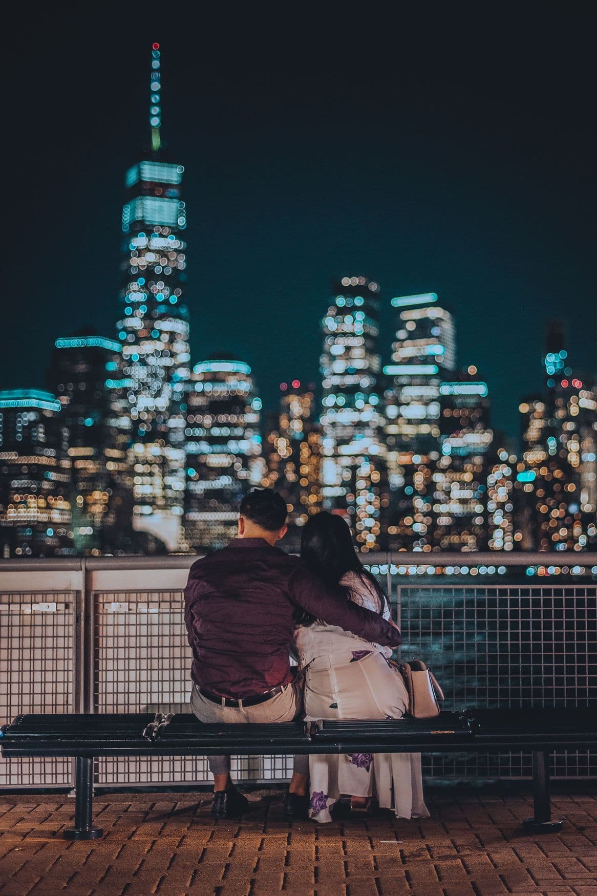 A couple on a bench looking at the Manhattan skyline at night.